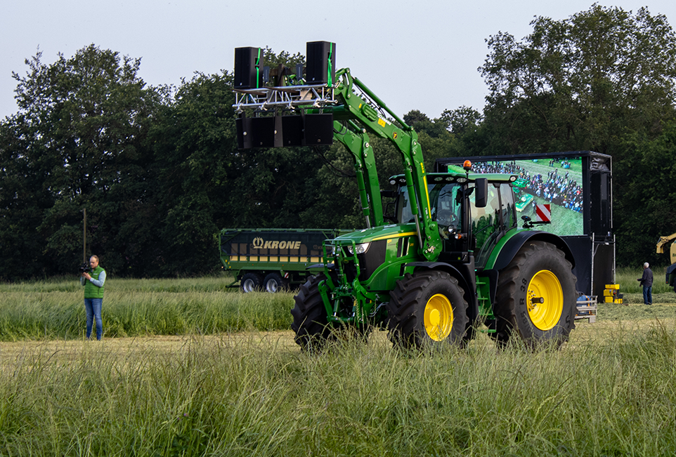 John Deere Traktor mit Frontlader an dem Lautsprecherboxen angebracht sind. Dahinter ein Krone Ladewagen und eine große Leinwand auf der ein Bild übertragen wird.